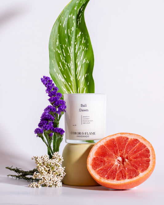 Color & Flame Handmade Bali Dawn candle styled with grapefruit, purple flowers, white botanicals, and a green leaf.
