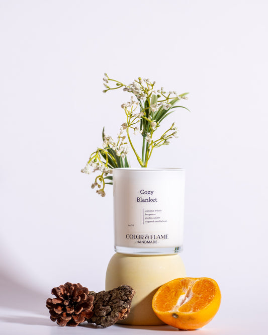 Color & Flame Handmade Cozy Blanket candle styled with an orange half, pine cone, wood, and delicate white flowers.