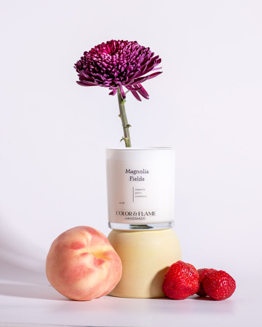 Color & Flame Handmade Magnolia Fields candle styled with a peach, strawberries, and a deep purple flower.