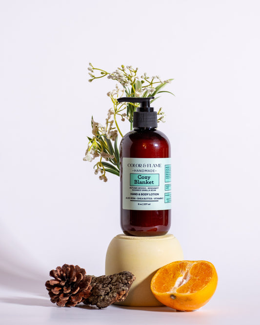 Color & Flame Handmade Cozy Blanket hand and body lotion styled with orange, pine cone, bark, and white flowers.
