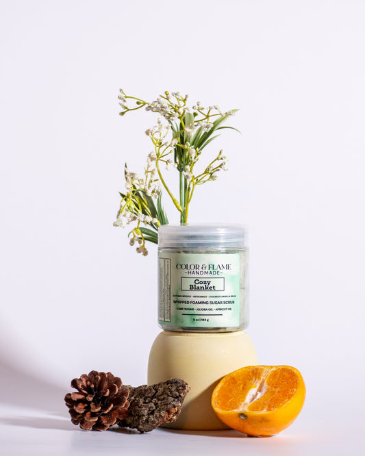 Color & Flame Handmade Cozy Blanket whipped foaming sugar scrub styled with evergreen, pine cone, bark, orange, and white flowers.
