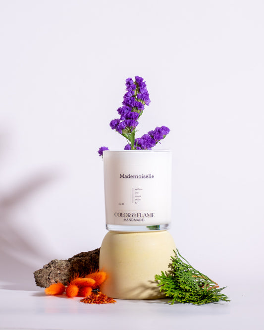 Color & Flame Handmade Mademoiselle candle styled with purple flowers, evergreen sprigs, bark, and warm dried botanicals.
