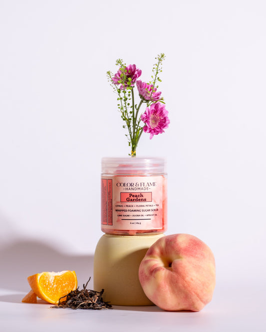 Color & Flame Handmade Peach Gardens whipped foaming sugar scrub styled with peach, orange, tea leaves, and pink flowers.
