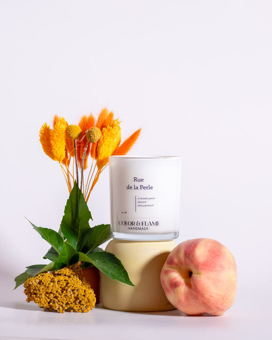 Color & Flame Handmade Rue de la Perle candle styled with a peach, green leaves, orange botanicals, and yellow dried flowers.
